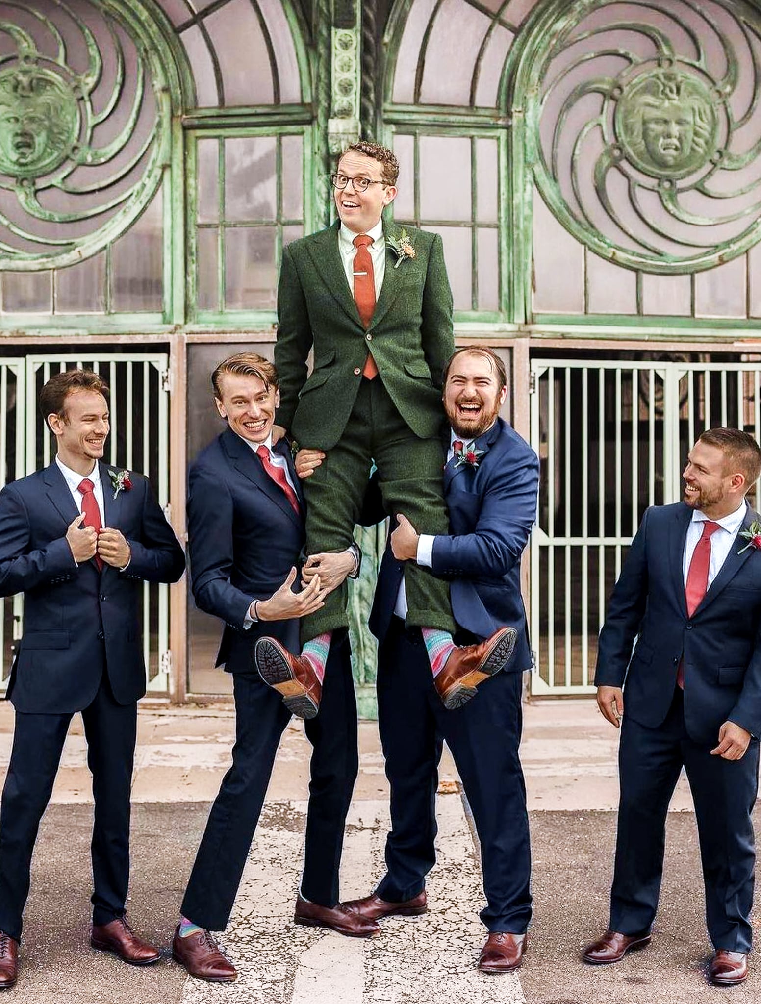 4 groomsmen lifting up groom on shoulders. Groom wearing green custom wedding suit. Groomsmen wearing navy blue suits.