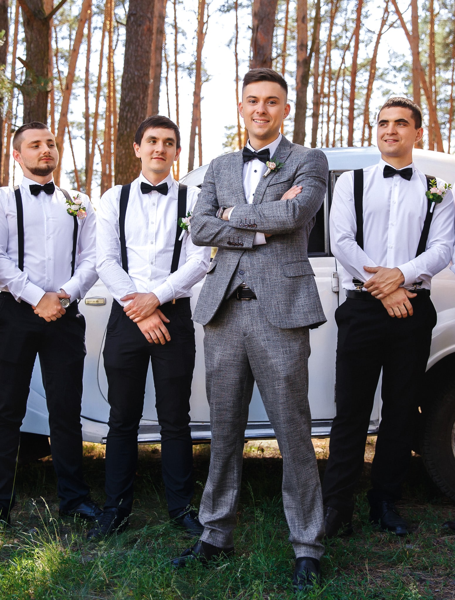 5 groomsmen with white shirts and black bowties without jackets on standing behind groom who is wearing a grey tweed custom wedding suit with a white tuxedo shirt and black bowtie.