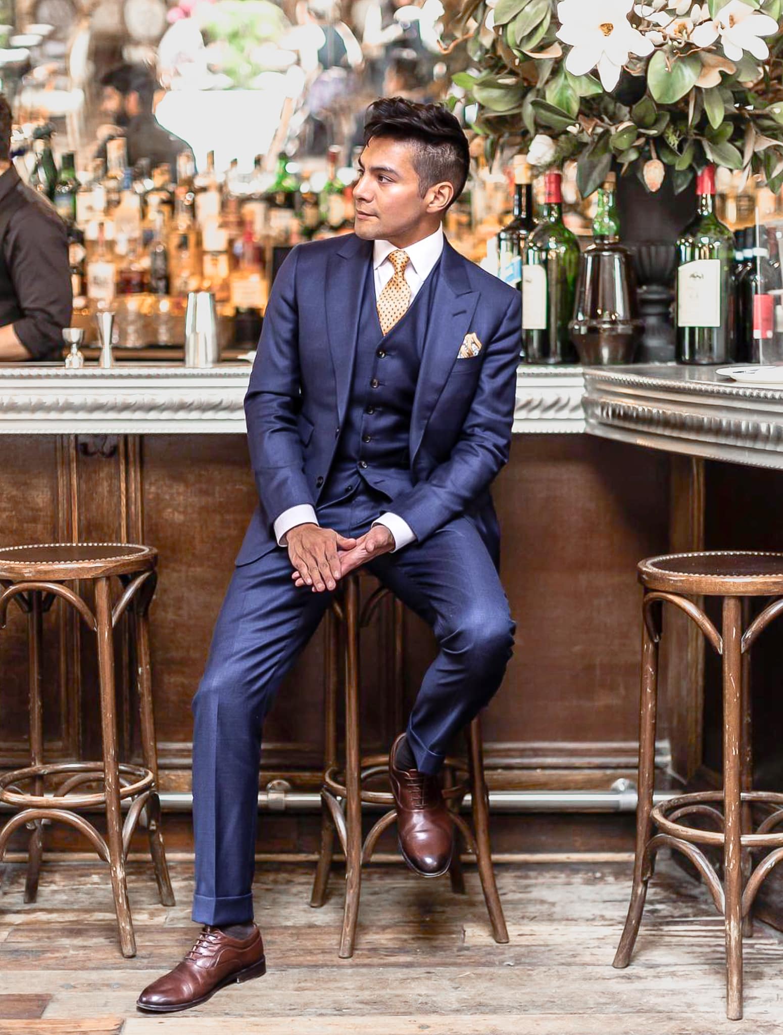 Groomsman sitting at bar wearing blue sharkskin suit solid with vest, white shirt and yellow tie.