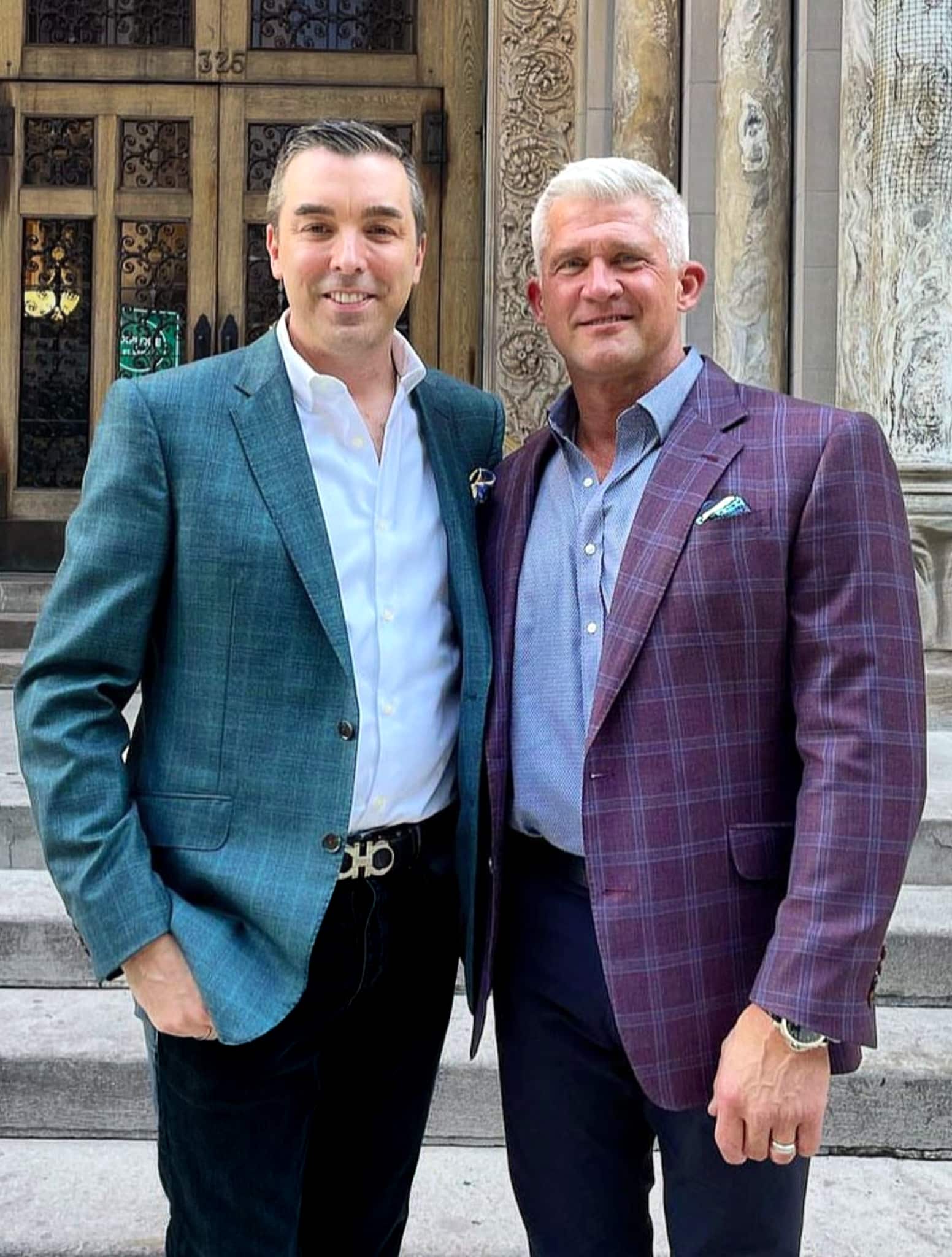 Groom and groom. One wearing red bespoke sportscoat with blue windowpane and one wearing blue bespoke sportscoat with tan windowpane. Both wearing navy trousers.