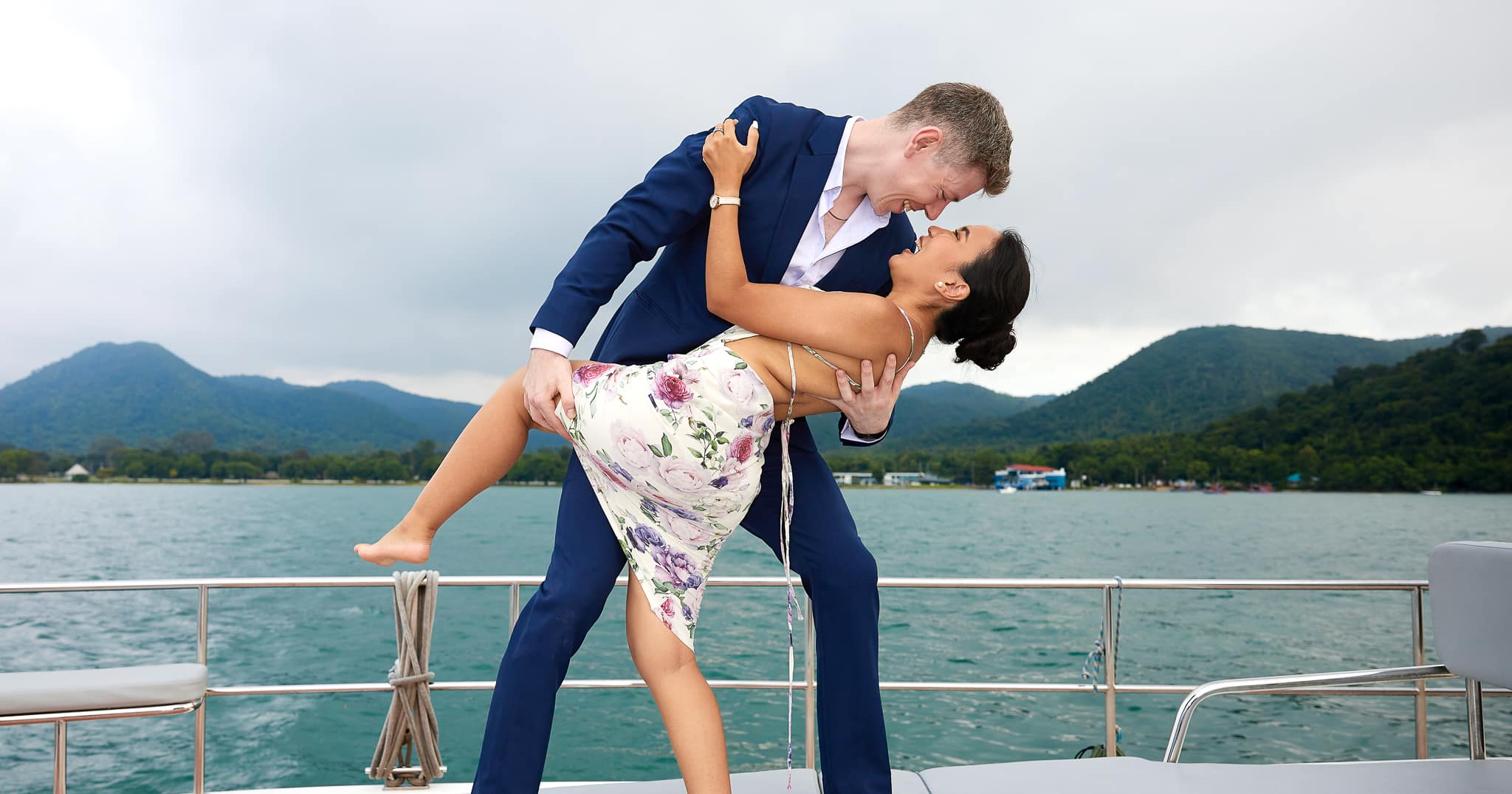 Groom on boat overlooking ocean dipping bride. Groom in navy blue bespoke suit with open collared blue shirt.