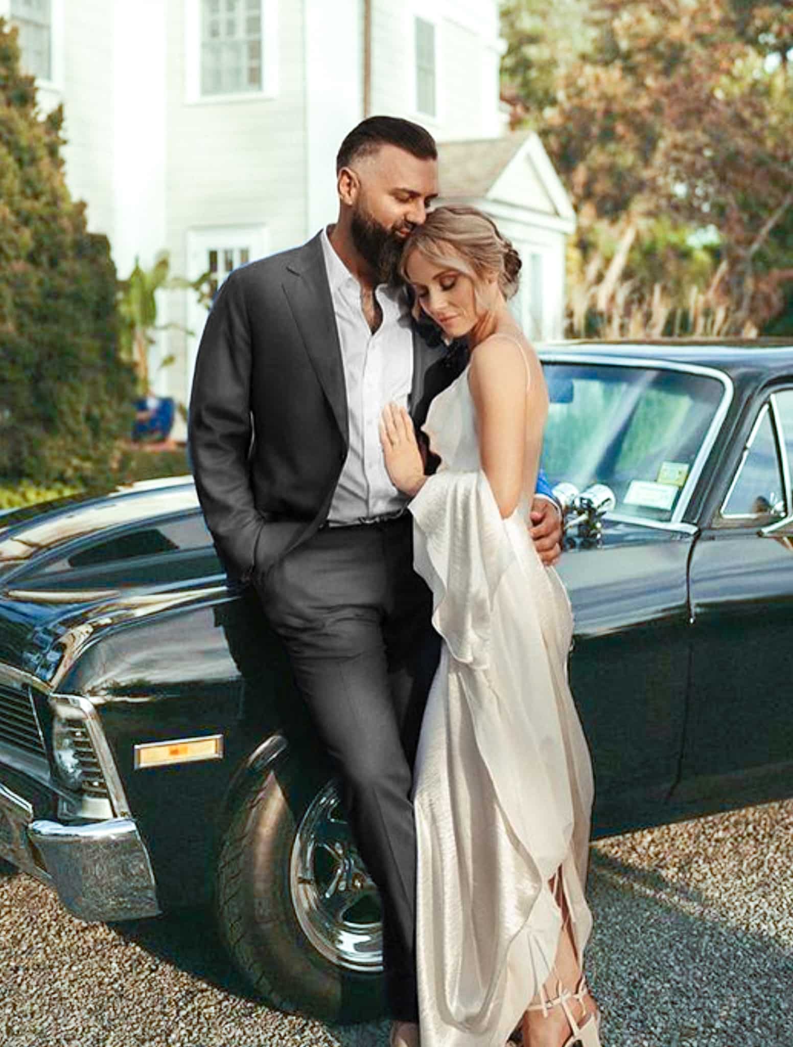 Bride and groom in front of antique car. Groom in Holland & Sherry solid black custom wedding suit.