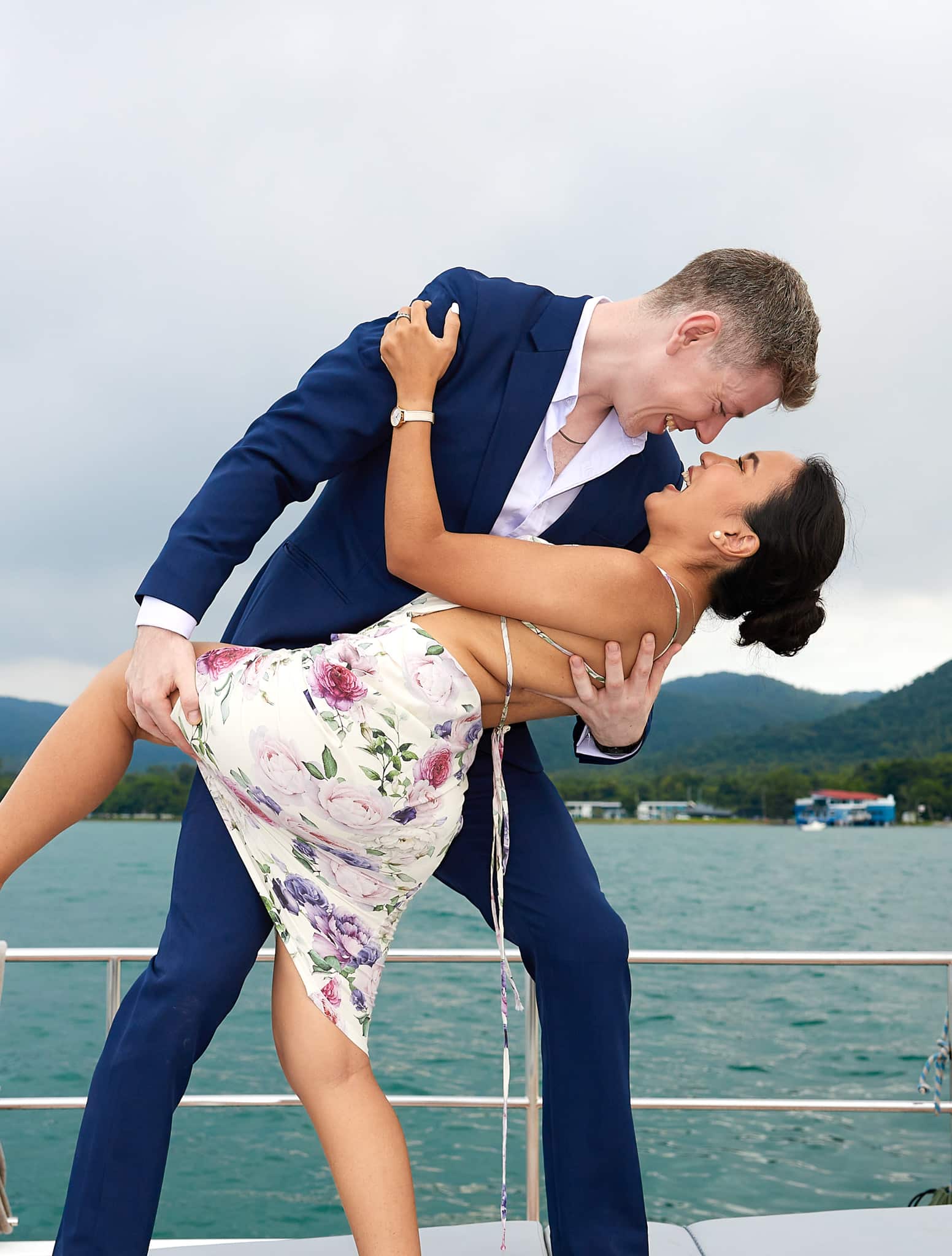 Groom on boat overlooking ocean dipping bride. Groom in navy blue bespoke suit with open collared blue shirt.